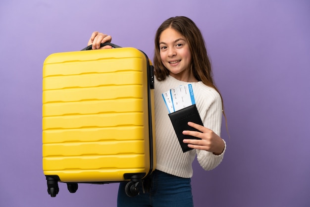 Mała kaukaska dziewczynka na fioletowym tle na wakacjach z walizką i paszportem