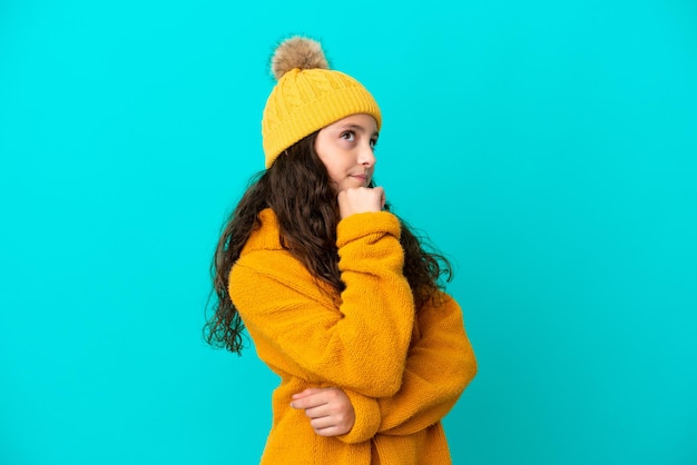 Mała kaukaska dziewczynka nosi czapkę zimową na białym tle na niebieskim tle patrząc z boku