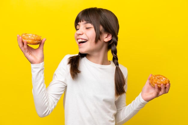 Mała kaukaska dziewczynka odizolowana na żółtym tle trzymająca pączki ze szczęśliwym wyrazem twarzy