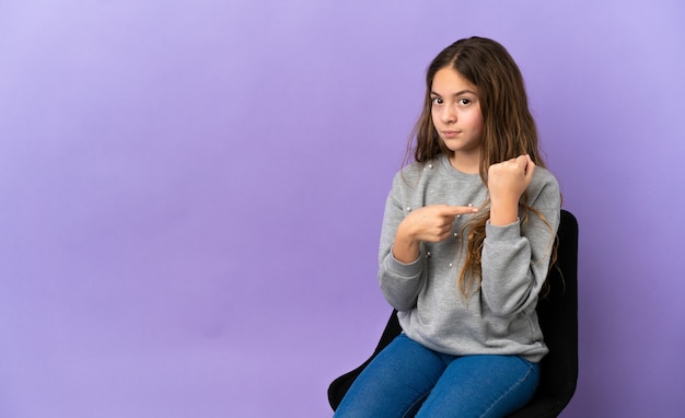 Mała kaukaska dziewczynka siedzi na krześle na fioletowym tle, robiąc gest spóźnienia