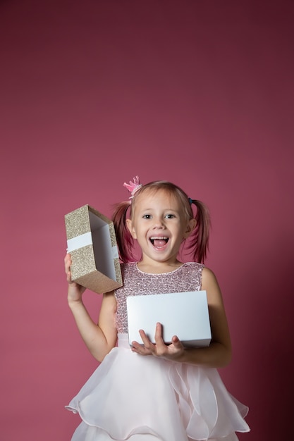 Mała kaukaska dziewczynka w świątecznej sukience z cekinami pozuje otwierając pudełko na różowym tle patrząc w kamerę