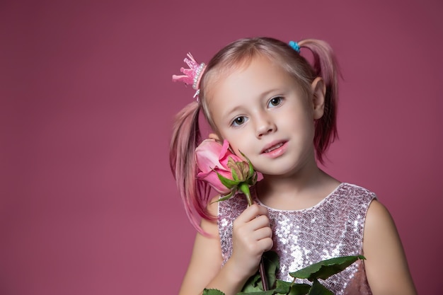 Mała kaukaska dziewczynka w świątecznej sukience z cekinami pozuje z kwiatem róży na różowym tle patrząc w kamerę