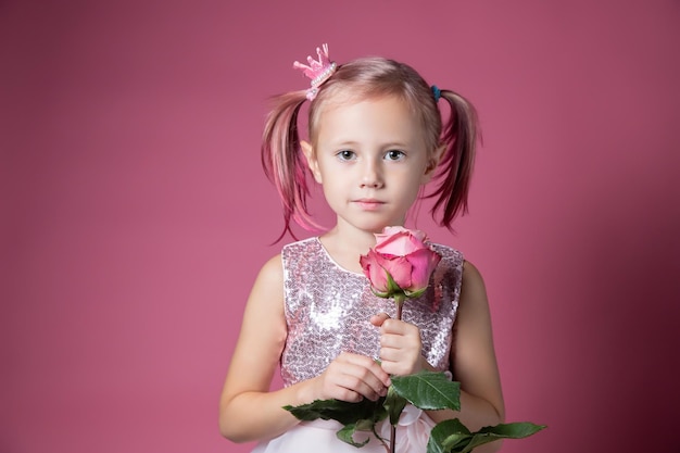 Mała kaukaska dziewczynka w świątecznej sukience z cekinami pozuje z kwiatem róży na różowym tle patrząc w kamerę