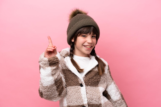 Mała kaukaska dziewczynka w zimowej kurtce na różowym tle pokazująca i unosząca palec na znak najlepszych