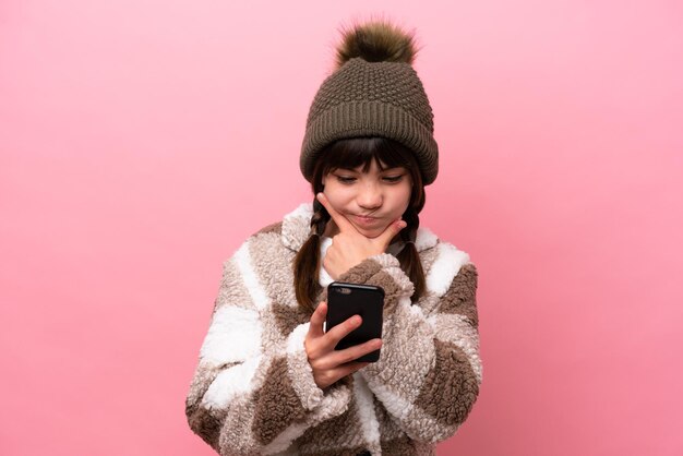Mała kaukaska dziewczynka w zimowej kurtce odizolowana na różowym tle myśląca i wysyłająca wiadomość