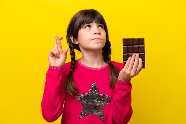 Mała kaukaska dziewczynka z czekoladą odizolowaną na żółtym tle z palcami skrzyżowanymi i życzącymi wszystkiego najlepszego