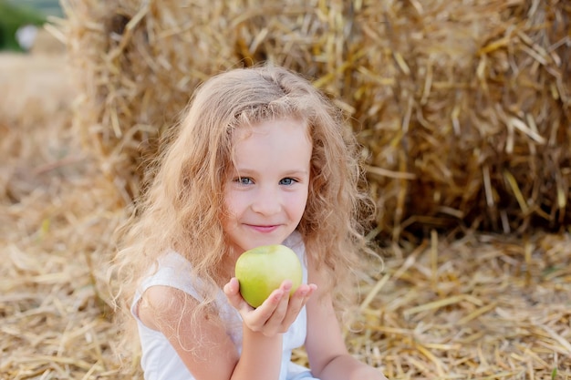 mała kręcona dziewczynka siedzi przy stogu siana na polu z zielonym jabłkiem w dłoniach