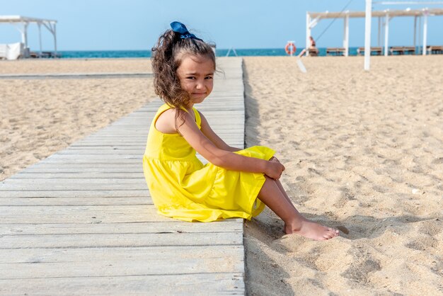 Mała kręcona dziewczynka w żółtej sukience siedzi na piaszczystej bezludnej plaży w słoneczny dzień zabawy wakacje