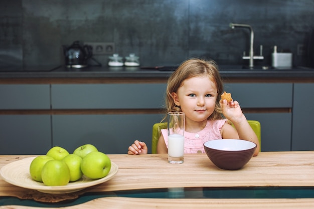 Mała piękna szczęśliwa dziewczynka w domu w kuchni przy stole z jabłkami i mlekiem