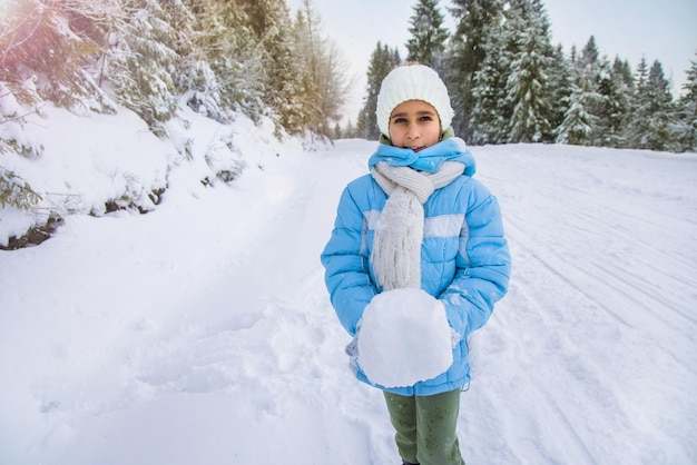 Mała przebiegła, wesoła dziewczyna w błękitnej kurtce i białej czapce z dzianiny stoi na zaśnieżonej drodze w sosnowym cichym zimowym lesie i trzyma dużą zimną śnieżkę