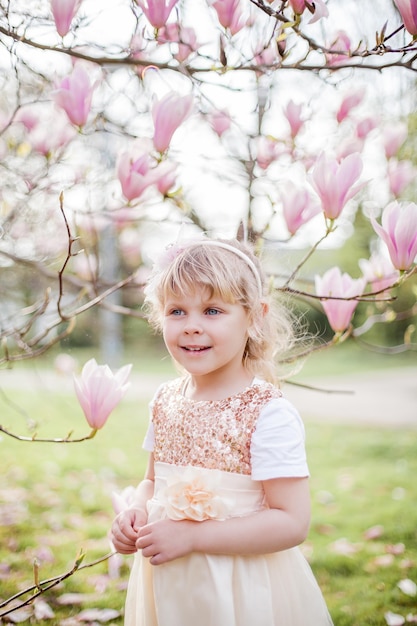 Mała śliczna blondynka 3 lata bawi się w parku w pobliżu kwitnącej magnolii. Wiosna.