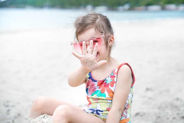 Mała śliczna dziewczyna w okularach bawi się w piasku na plaży nad morzem.