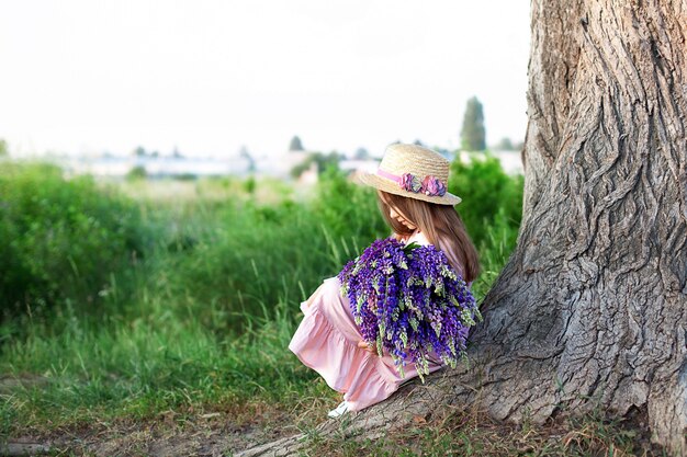 mała śliczna dziewczyna w słomkowym kapeluszu siedzi na drzewie z ogromnym bukietem łubinów polnych kwiatów. pojęcie natury i romansu.