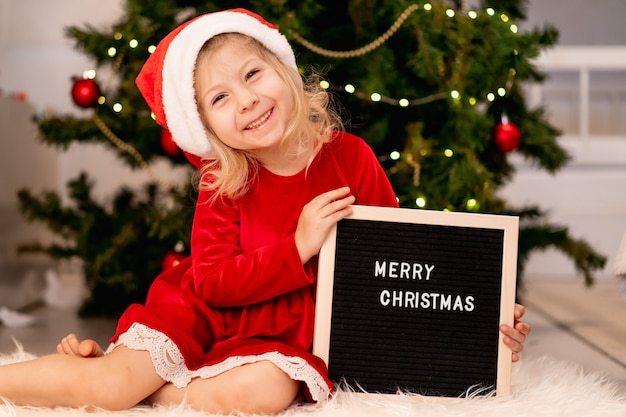 Mała śliczna dziewczynka w czapce Świętego Mikołaja i czerwonej świątecznej sukience leży pod choinką i śmieje się obok czarnej tablicy i białych liter szczęśliwego nowego roku 2021. Zdjęcie wysokiej jakości
