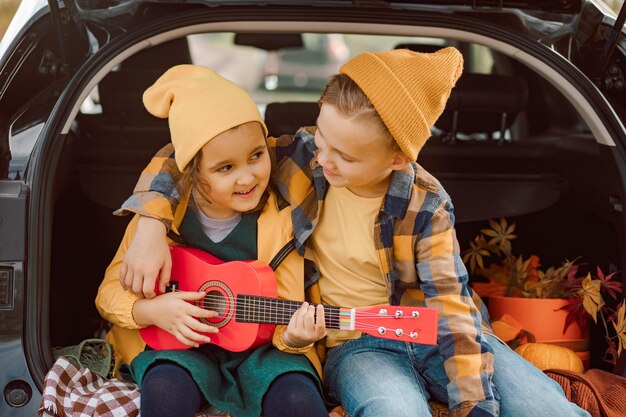 Zdjęcie mała śliczna uśmiechnięta dziewczyna i chłopiec stojący w pobliżu na otwartym bagażniku samochodu dziewczyna gra na małej gitarze dziecko odpoczywa z rodziną w naturze sezon jesienny