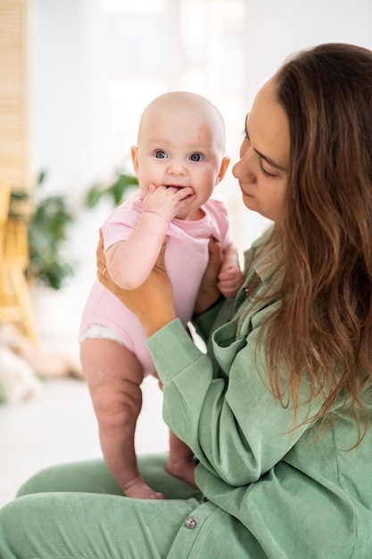 Mała śliczna uśmiechnięta dziewczynka w różowym body w ramionach matki w sypialni w domu uściski mamy i córki opieka nad dzieckiem opieka mamy