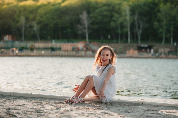 Mała słodka dziewczyna w białej sukience siedzi na piasku na plaży w ciepłych kolorach latem