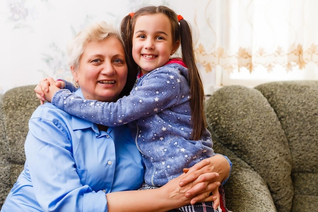 Mała słodka dziewczynka i jej babcia spędzają razem czas w domu. Zabawa, przytulanie i uśmiechanie się siedząc na kanapie.