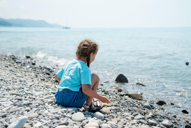 Mała słodka dziewczynka siedzi na kamienistej plaży i bawi się kamykami