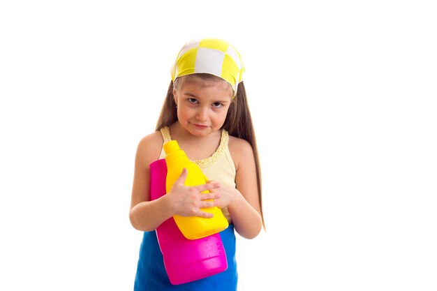 Mała smutna dziewczynka w niebieskim fartuchu z żółtą chustką trzymająca żółte i różowe butelki w studio