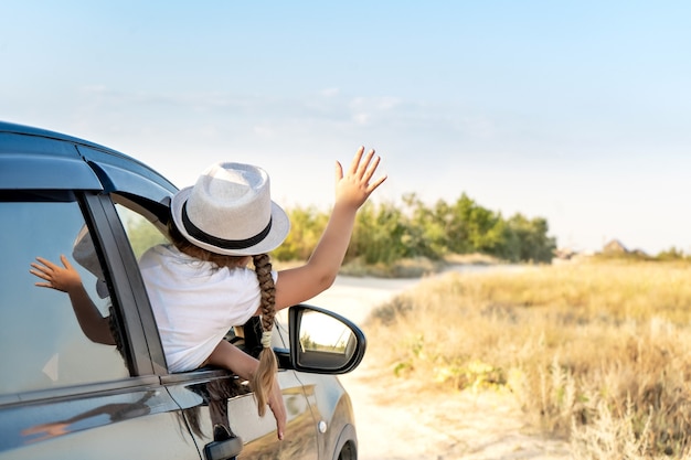 Mała szczęśliwa dziewczynka w kapeluszu podnosi rękę z okna samochodu SUV z gnaturem na tleSamochód podróżny