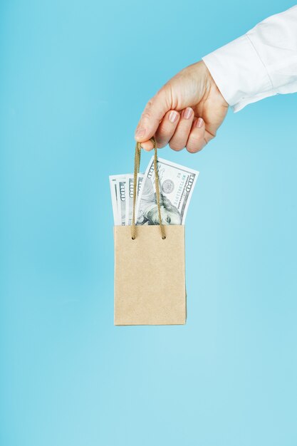 Mała torba pomocnicza wykonana z papieru w wyciągniętej dłoni z dolarami amerykańskimi na niebieskim tle. Układ szablonu opakowania z miejscem na kopiowanie, reklamę.