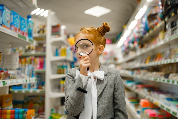 Zdjęcie mała uczennica patrzy przez szkło powiększające, robi zakupy w sklepie papierniczym