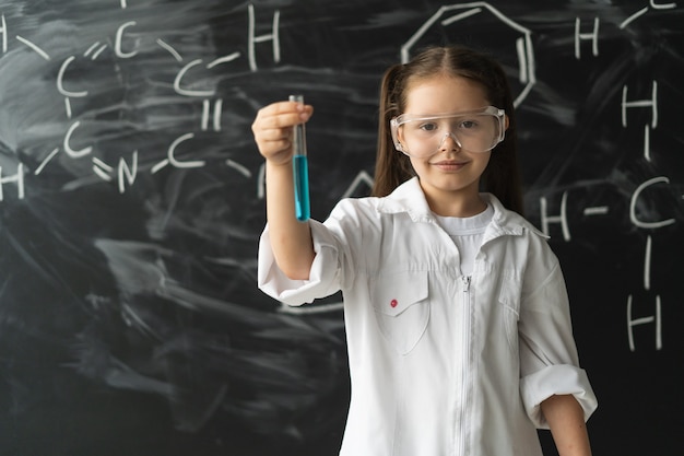 Mała uczennica w okularach i białym fartuchu stoi obok tablicy testowej lekcji chemii ...