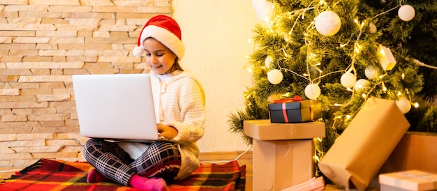 Mała urocza dziewczynka w czapce Mikołaja komunikuje się online z krewnymi na laptopie podczas kwarantanny Covid-19, dystansu społecznego, samoizolacji, dzieci zostały w domu podczas świąt Bożego Narodzenia..