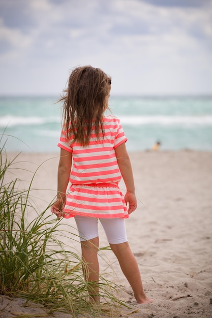 Mała urocza dziewczynka w pasiastej sukience stojąca na piaszczystej plaży i patrząca na ocean