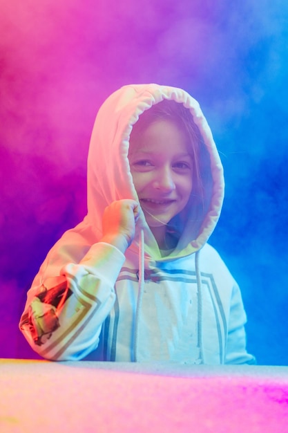 Mała uśmiechnięta dziewczyna w bluzie z kapturem stoi w kolorowym dymie i patrzy w kamerę