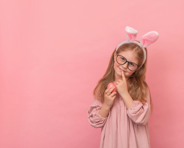 Mała uśmiechnięta dziewczyna w okularach z uszami królika i pisanką na różowym tle