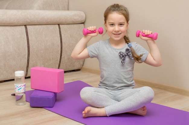 Mała uśmiechnięta dziewczynka siedzi na macie fitness i trzyma w dłoniach kettlebells