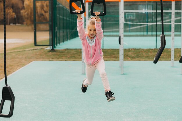 Mała wesoła dziewczynka bawi się na placu zabaw. Dziecko jest zaangażowane na kółkach gimnastycznych na ulicy. Dziecko uprawia sport na symulatorach.