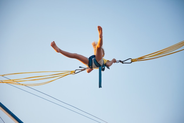 Mała Wesoła Dziewczynka Leci Na Sprężystych, Jasnych Gumkach I Skacze Na Trampolinie Ciesząc Się Długo Oczekiwanymi Wakacjami W Ciepłym Słońcu