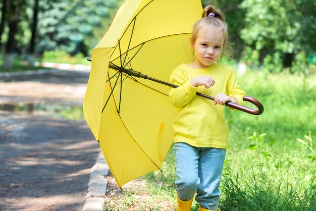 Zdjęcie mała wesoła dziewczynka spaceruje po deszczu w kaloszach i jesienią pod żółtym parasolem