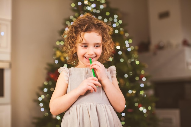 Mała wesoła dziewczynka z kręconymi włosami stoi w salonie z ozdobioną choinką na tle marzy o prezentach trzymając w ustach zielony ołówek Świąteczna koncepcja nowego roku