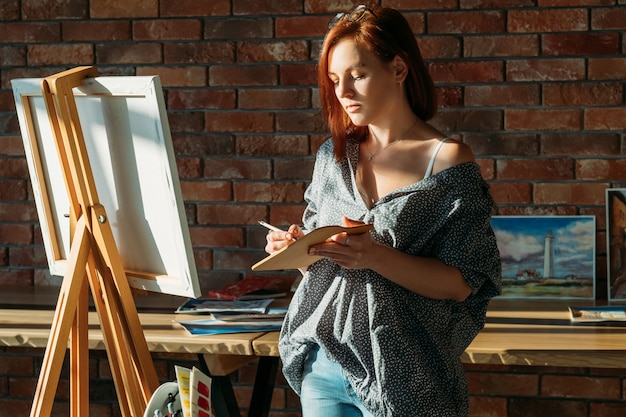 Malarstwo artystyczne. Obszar roboczy Studio. Ruda kobieta ze sztalugą, mieszanie kolorów na drewnianej palecie.