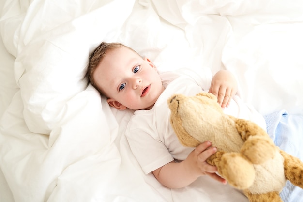 Małe dziecko o niebieskich oczach trzyma zabawkę leżącą na łóżku. Widok z góry.