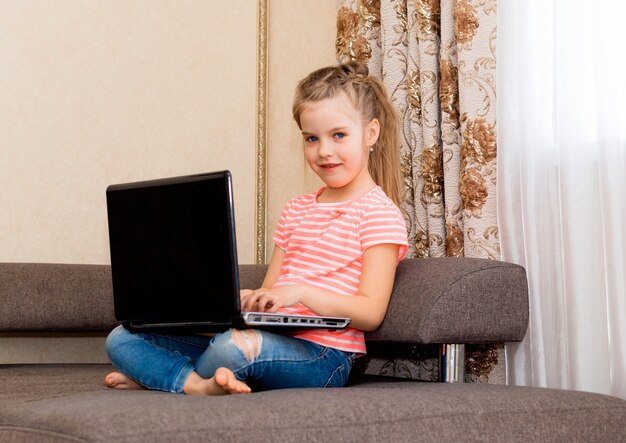 Małe dziecko surfuje po Internecie siedząc na szarej kanapie. dziewczyna z laptopem na kanapie siedzi