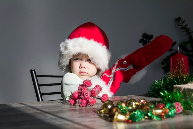 Małe dziecko w czapce Świętego Mikołaja i czerwonych rękawiczkach ze świątecznymi prezentami siedzi przy noworocznym stole