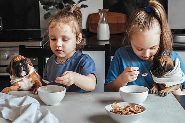 małe dziewczynki w kuchni karmią śniadanie małym zabawnym szczeniakom