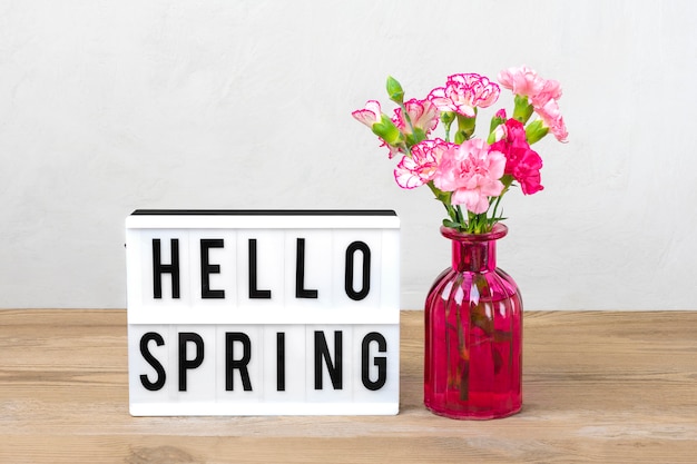 Małe Kolorowe Różowe Goździki W Wazonie, Lightbox Z Tekstem Hello Spring, Flamingowa Figura Na Drewnianym Stole I Szara ściana. Witaj Wiosno