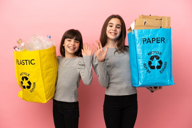 Małe siostry przetwarzają papier i plastik na białym tle na różowym tle pozdrawiając ręką ze szczęśliwym wyrazem twarzy