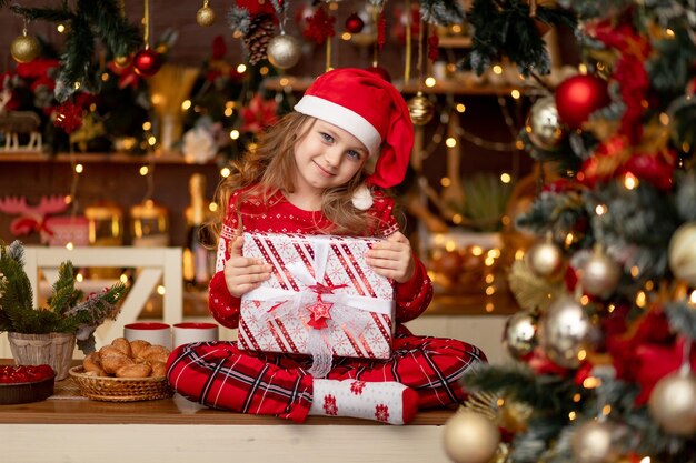 Zdjęcie małe słodkie dziecko dziewczynka w czerwonym swetrze i czapce świętego mikołaja w ciemnej kuchni z choinką raduje się prezentami i czeka na nowy rok lub boże narodzenie