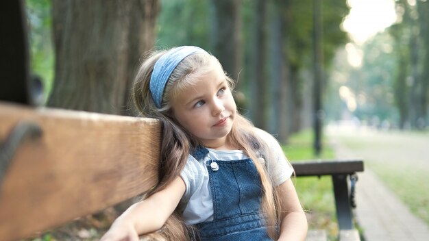 Małe szczęśliwe dziecko dziewczynka siedzi na ławce w parku latem.