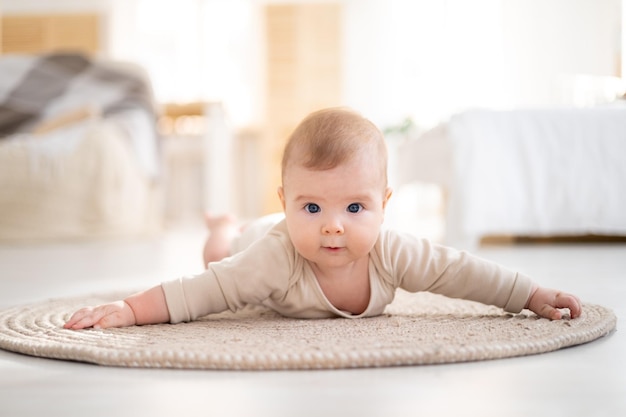 Małe zdrowe dziecko w bawełnianym body leży na brzuchu na dywanie w jasnym salonie domu patrzy w kamerę uśmiecha się