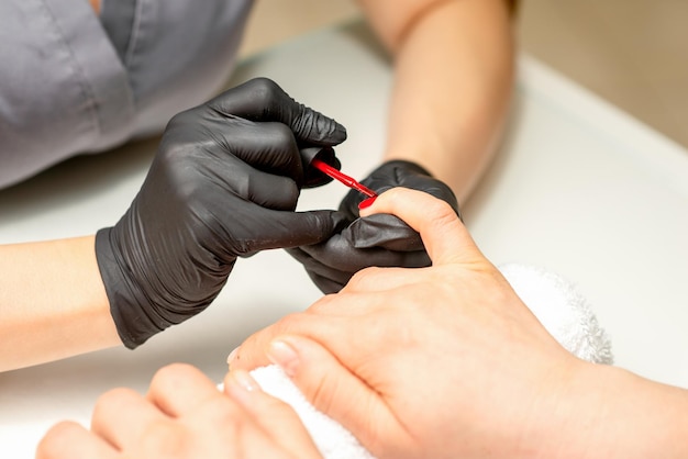 Malowanie lakierem do manicure Zbliżenie mistrza manicure w gumowych czarnych rękawiczkach nakładających czerwony lakier na kobiecy paznokieć w salonie kosmetycznym