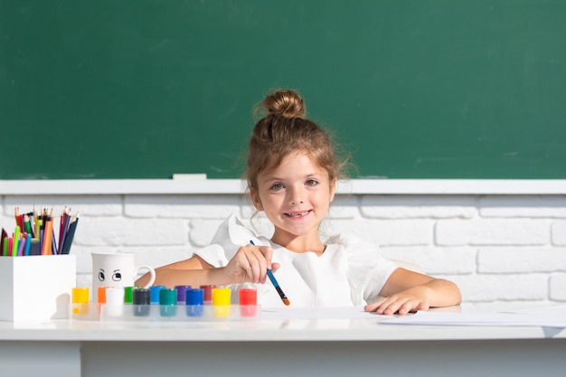 Malowanie lekcja szkolna rysowanie sztuki Dziecko dziewczynka rysuje w klasie, siedząc przy stole, bawiąc się na tle szkolnej tablicy
