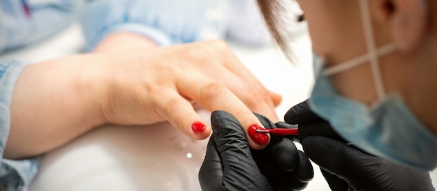 Malowanie paznokci kobiety. Ręce manikiurzystki w czarnych rękawiczkach, stosując czerwony lakier do paznokci na kobiece paznokcie w salonie piękności.